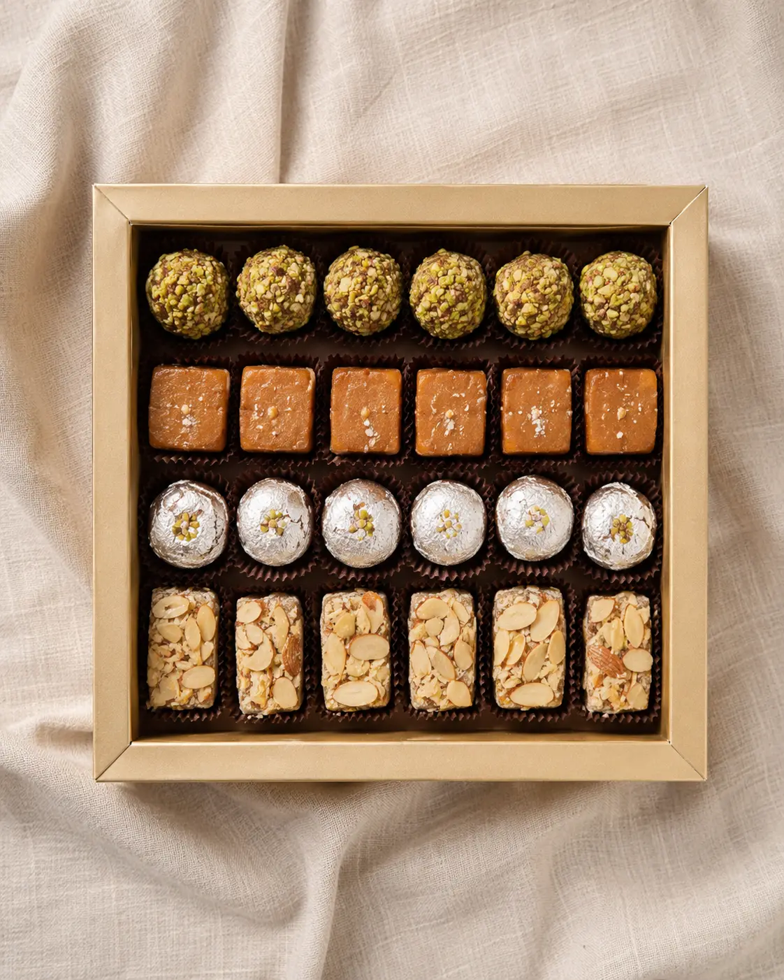 A shallow gold tray of Indian sweets shot from directly above on linen, four rows of confectionery in it and no lid on the tray