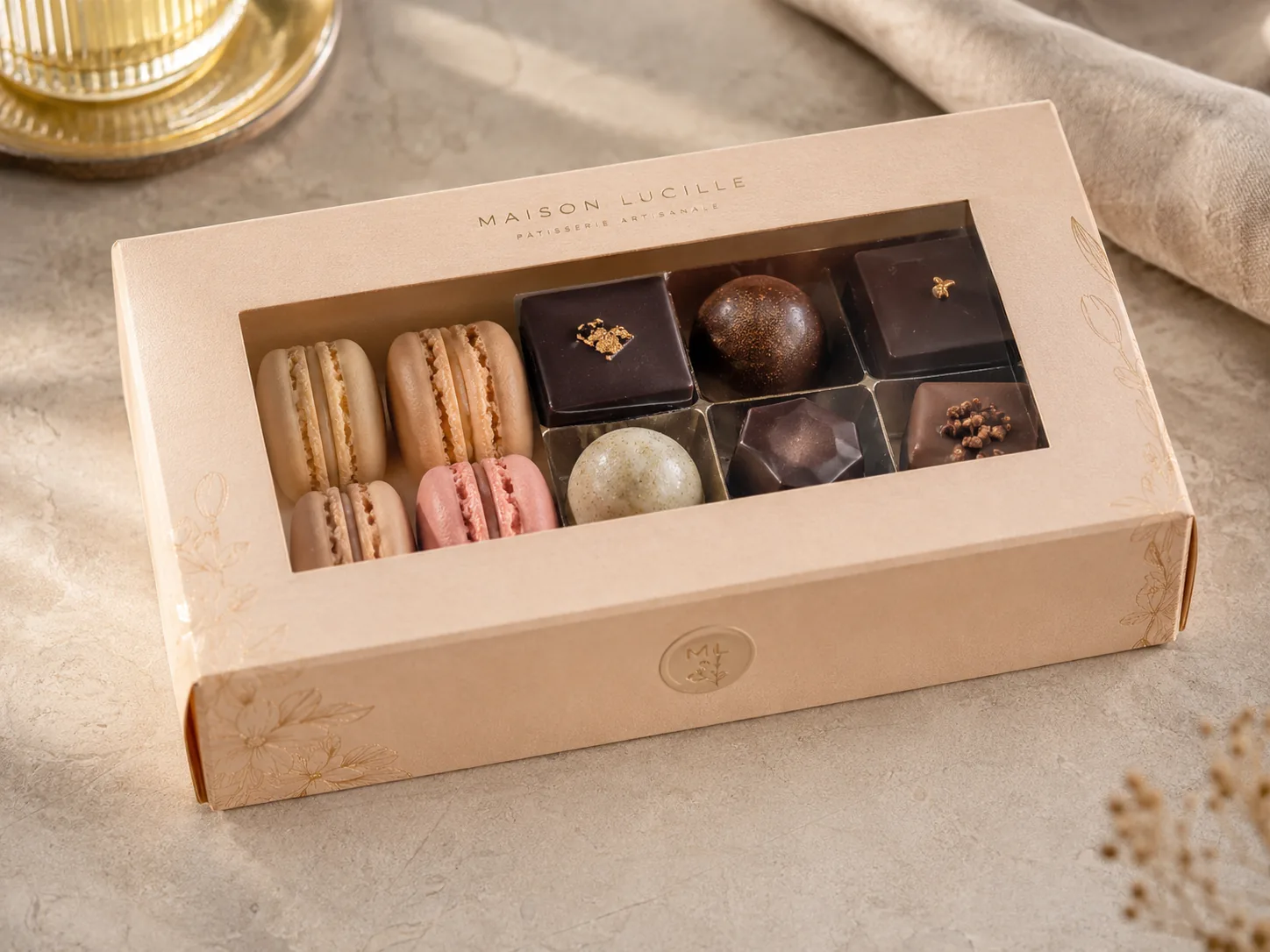 A pale blush window carton of macarons and chocolates seen from above, a clear patch across the large aperture cut into the top panel.