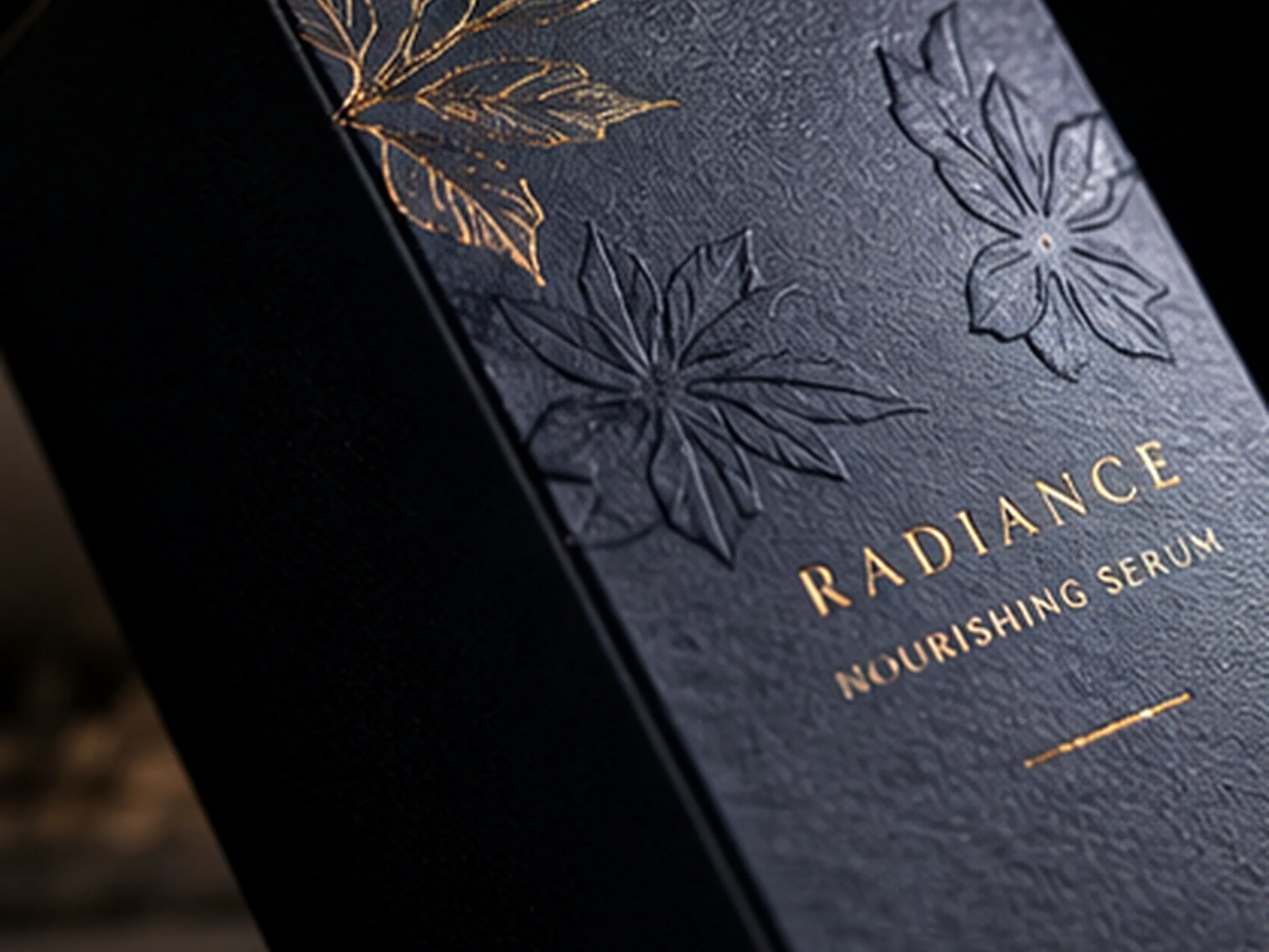 A navy soft touch serum carton with a hot foil gold leaf, blind embossed flowers and a gold foil wordmark under raking light