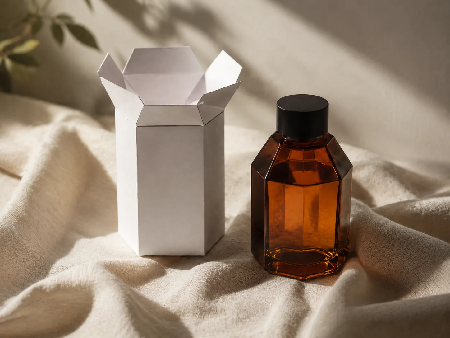 A white unprinted hexagonal carton standing on linen in daylight beside the faceted amber bottle it was drawn around, nothing branded in frame.