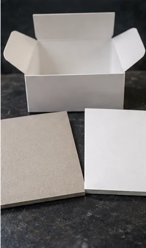 A gray back sheet and a white back sheet lying side by side on dark stone, a plain white carton standing open behind them with its flaps up