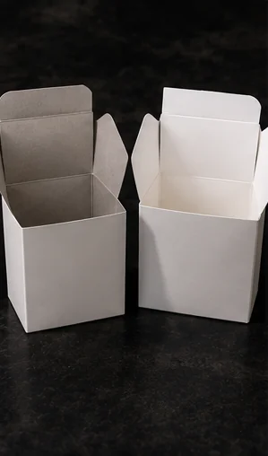 Two plain white cartons standing open on a dark stone surface, the one on the left gray inside and the one on the right white inside, their top flaps standing up
