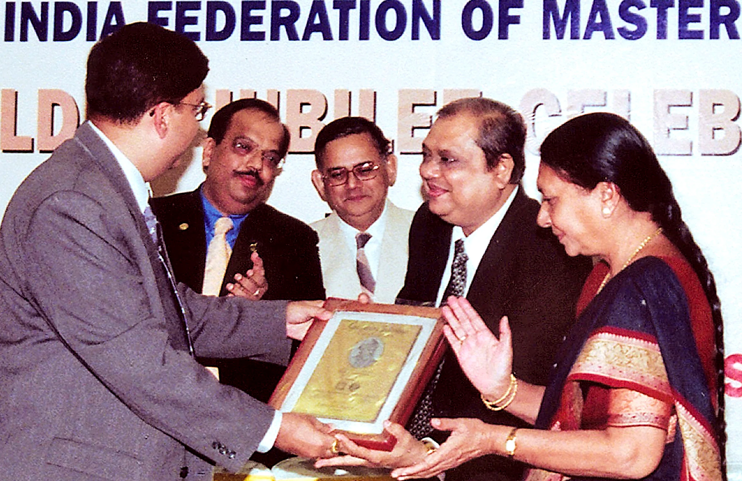 A framed plaque being presented on stage under an All India Federation of Master Printers Golden Jubilee banner, four people around it.