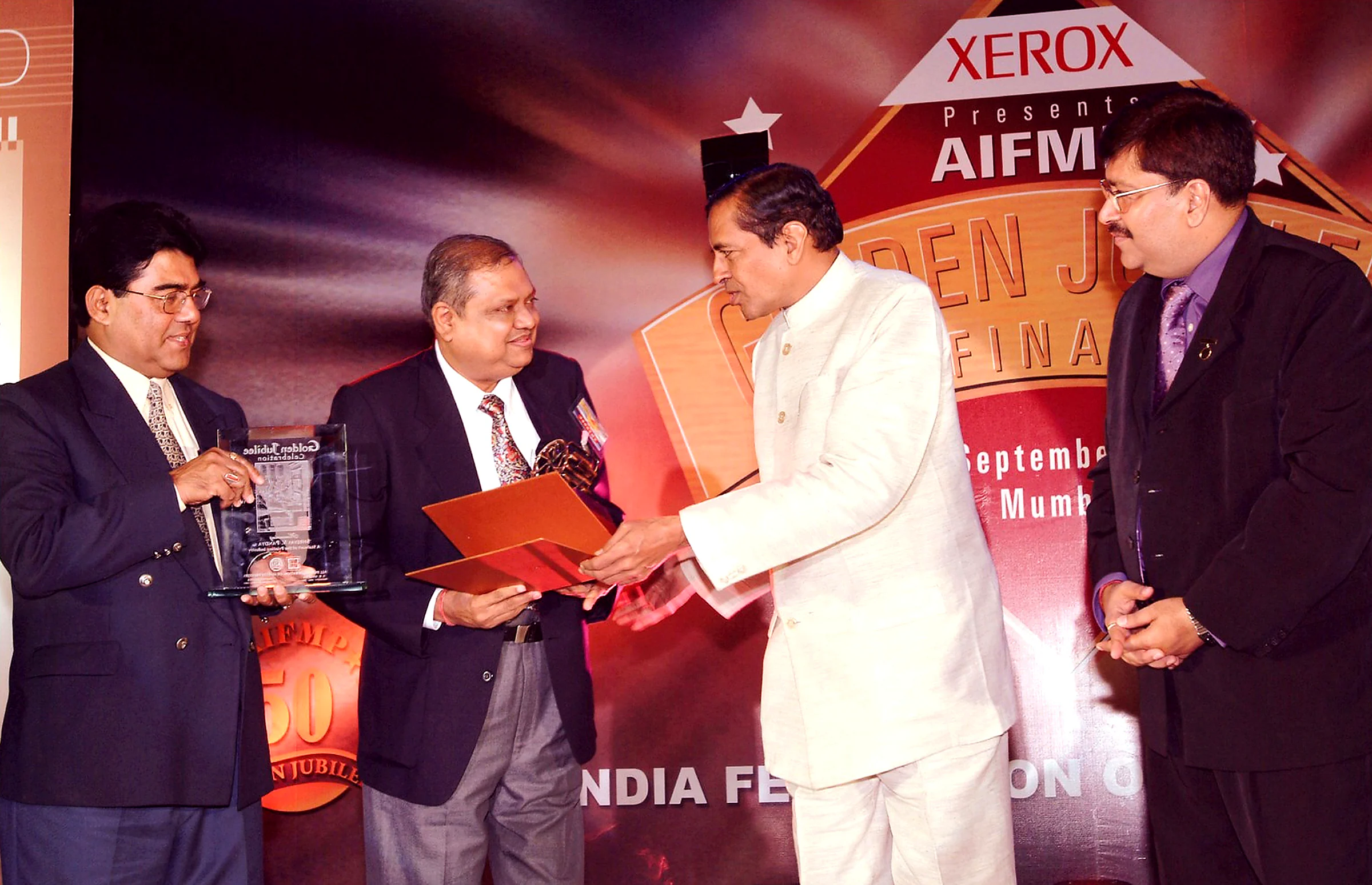 Shreyas Pandya receiving a certificate on stage at the AIFMP Golden Jubilee in Mumbai, a glass trophy reading A Stalwart of the Printing Industry held beside him.