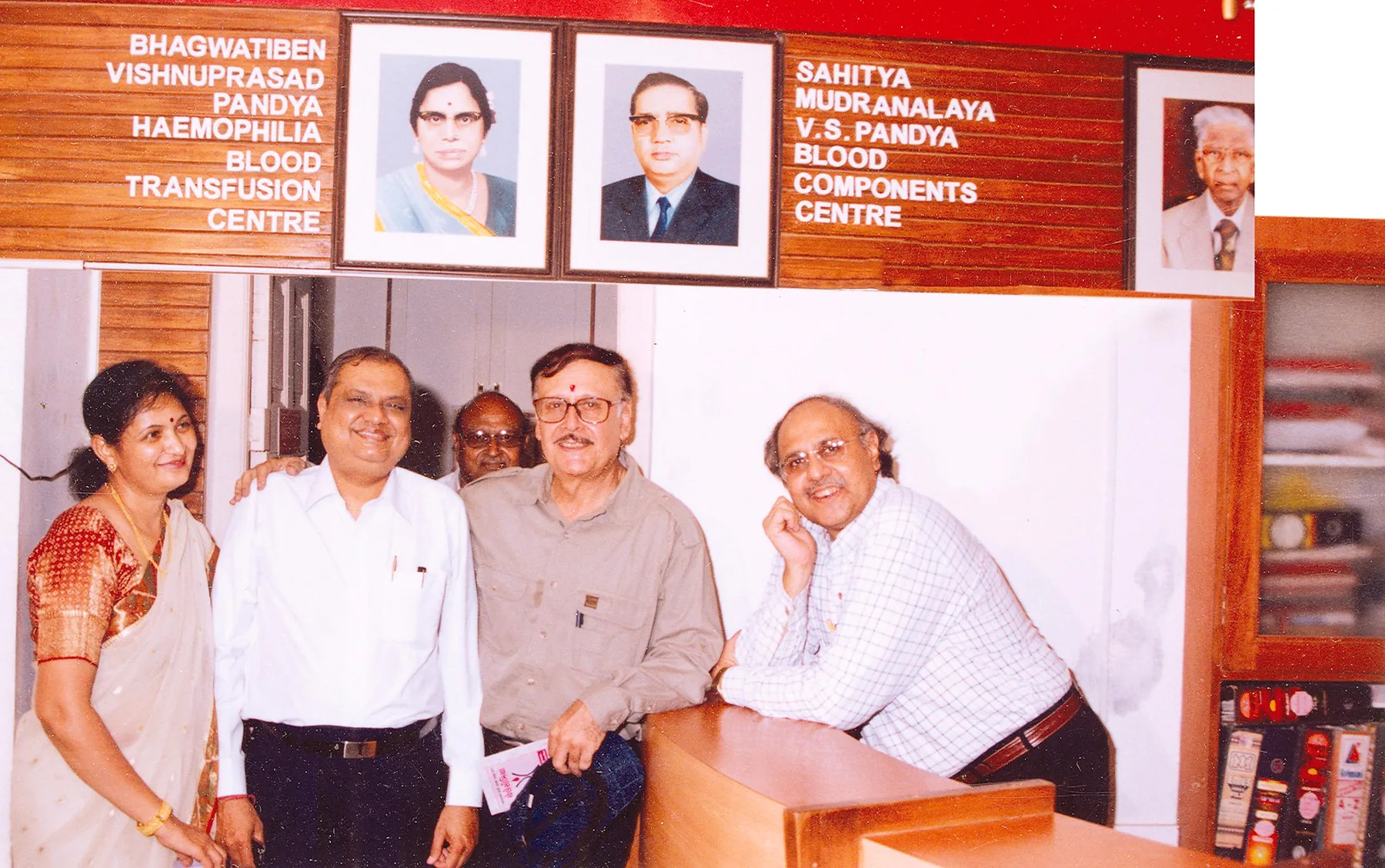 Five people standing under framed portraits and wooden signage naming the Sahitya Mudranalaya V.S. Pandya Blood Components Centre and the Bhagwatiben Vishnuprasad Pandya Haemophilia Blood Transfusion Centre.