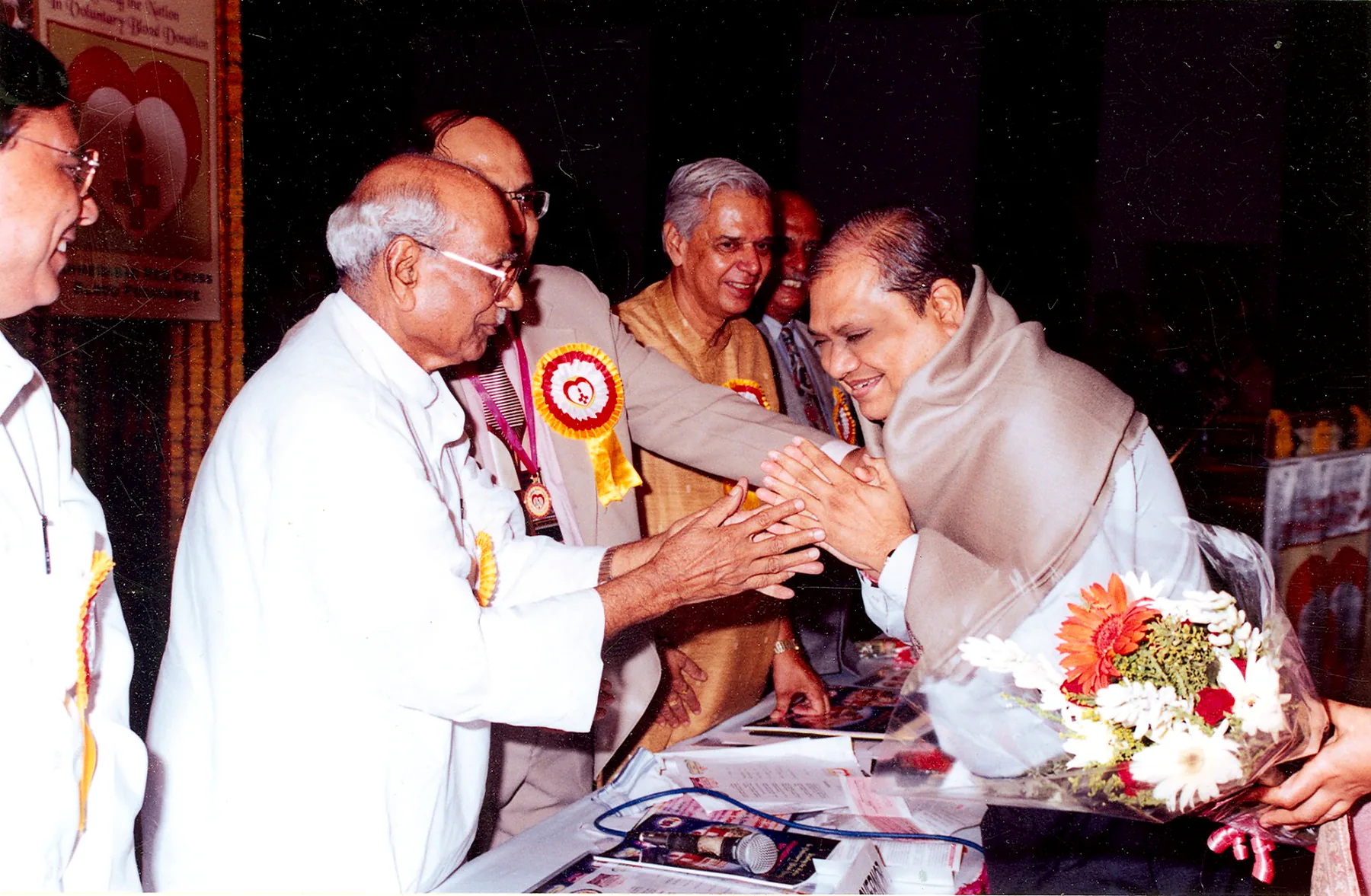 A man in a shawl, hands folded in greeting, being honoured on stage at a voluntary blood donation ceremony, a bouquet held beside him.