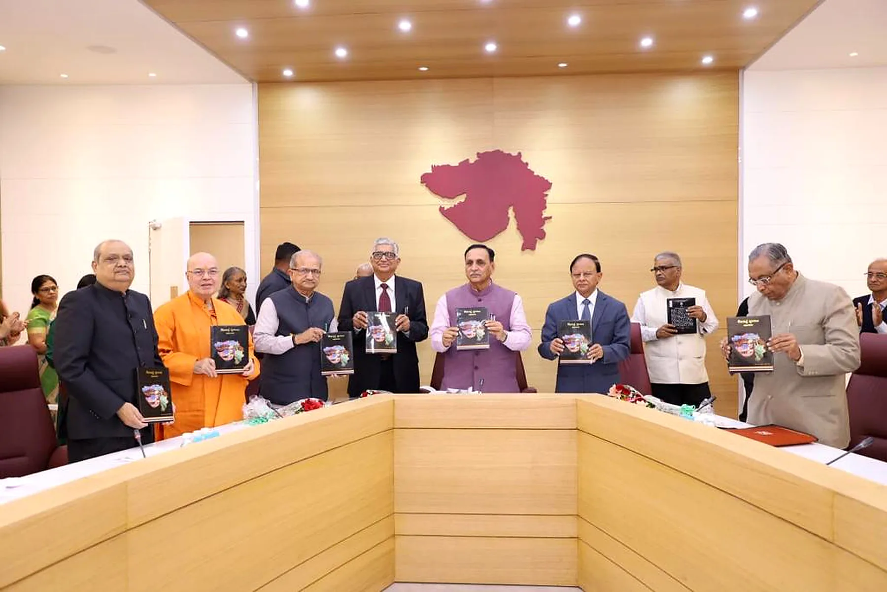Nine people standing behind a conference table holding up copies of the same book, a maroon map of Gujarat on the wood panelled wall behind them.