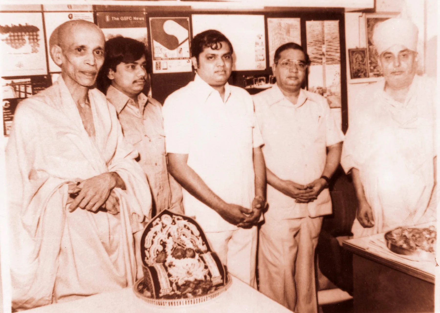 A sepia photograph of five men standing in a row in an office hung with printed posters, one of them from The GSFC News, an embroidered offering on the table in front of them.