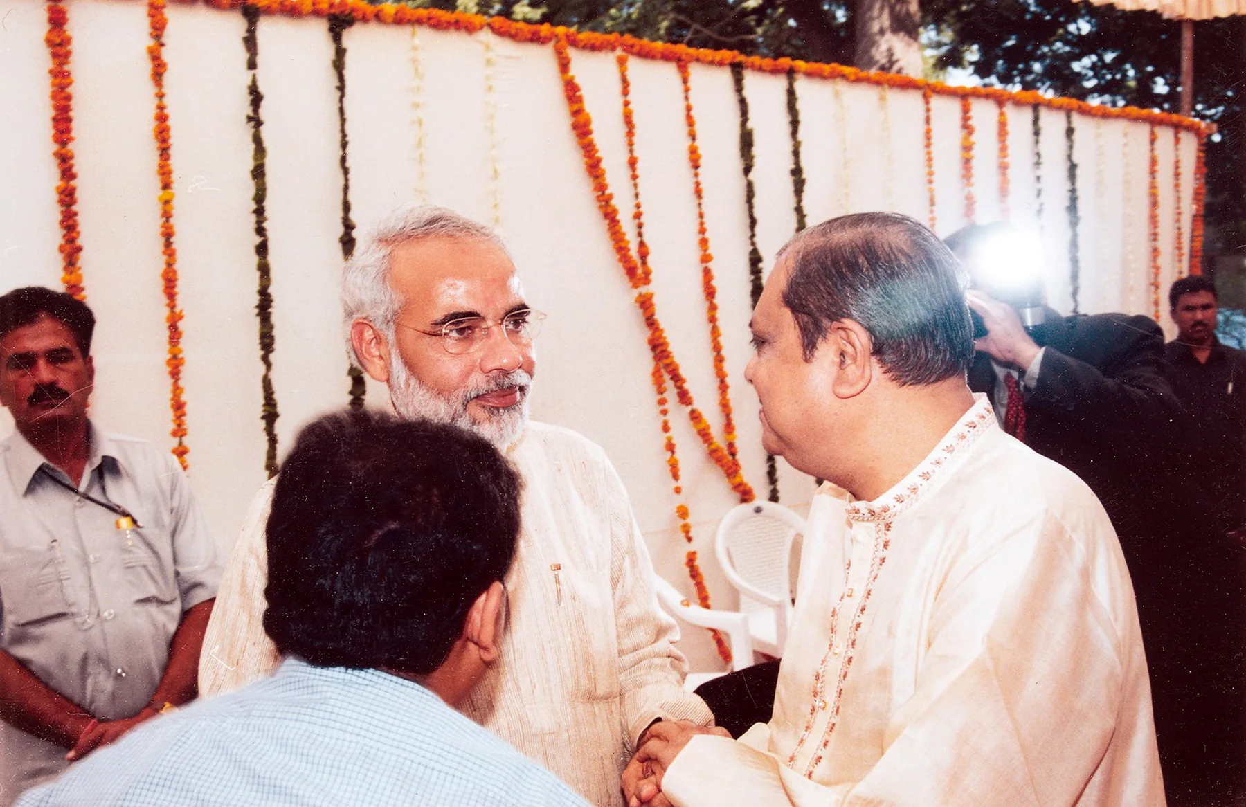 Two men in kurtas shaking hands in front of a white wall hung with marigold garlands, a photographer’s flash going off behind them.