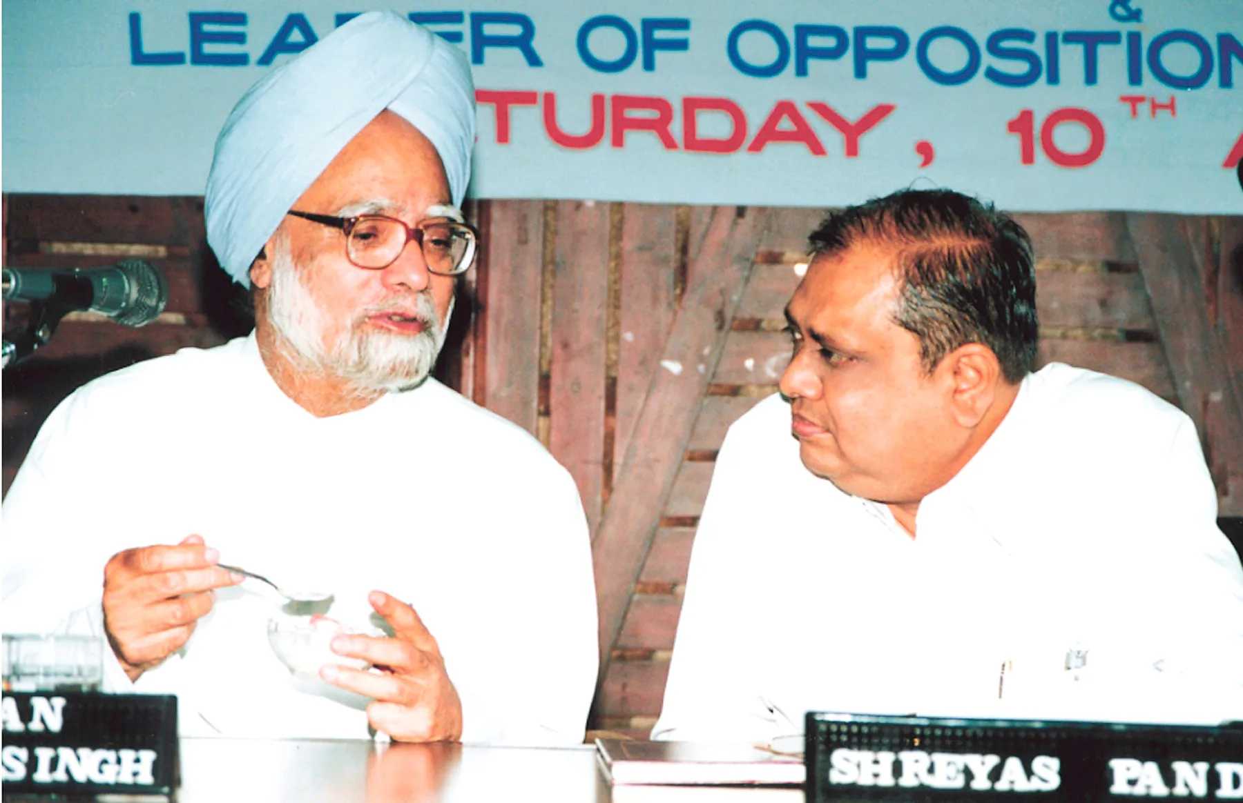 Two men in white at a head table under a banner reading Leader of Opposition, one behind a name plate reading Shreyas Pandya.