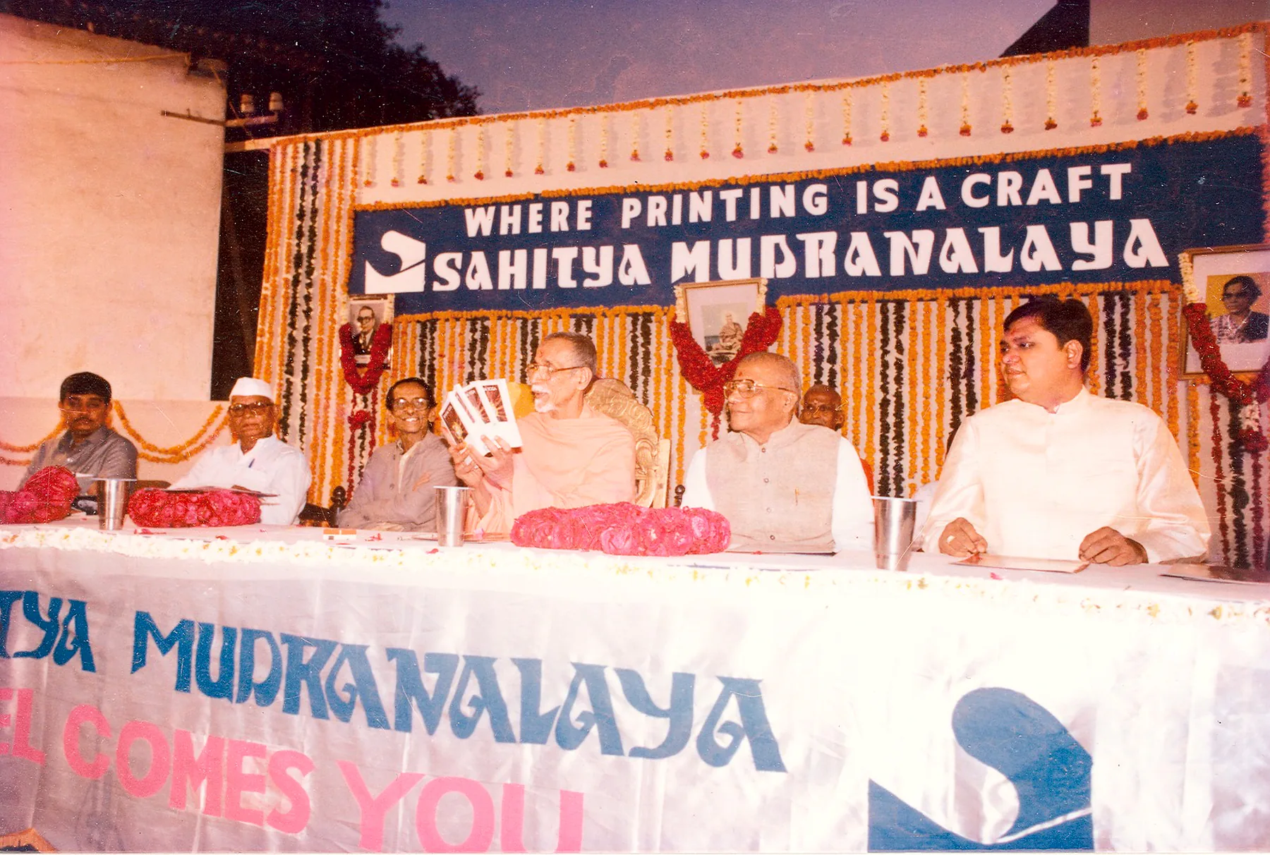 Guests seated at a long table on a stage hung with marigold garlands, under a banner reading Where printing is a craft, Sahitya Mudranalaya. A man in saffron robes at the center holds up a small printed booklet.