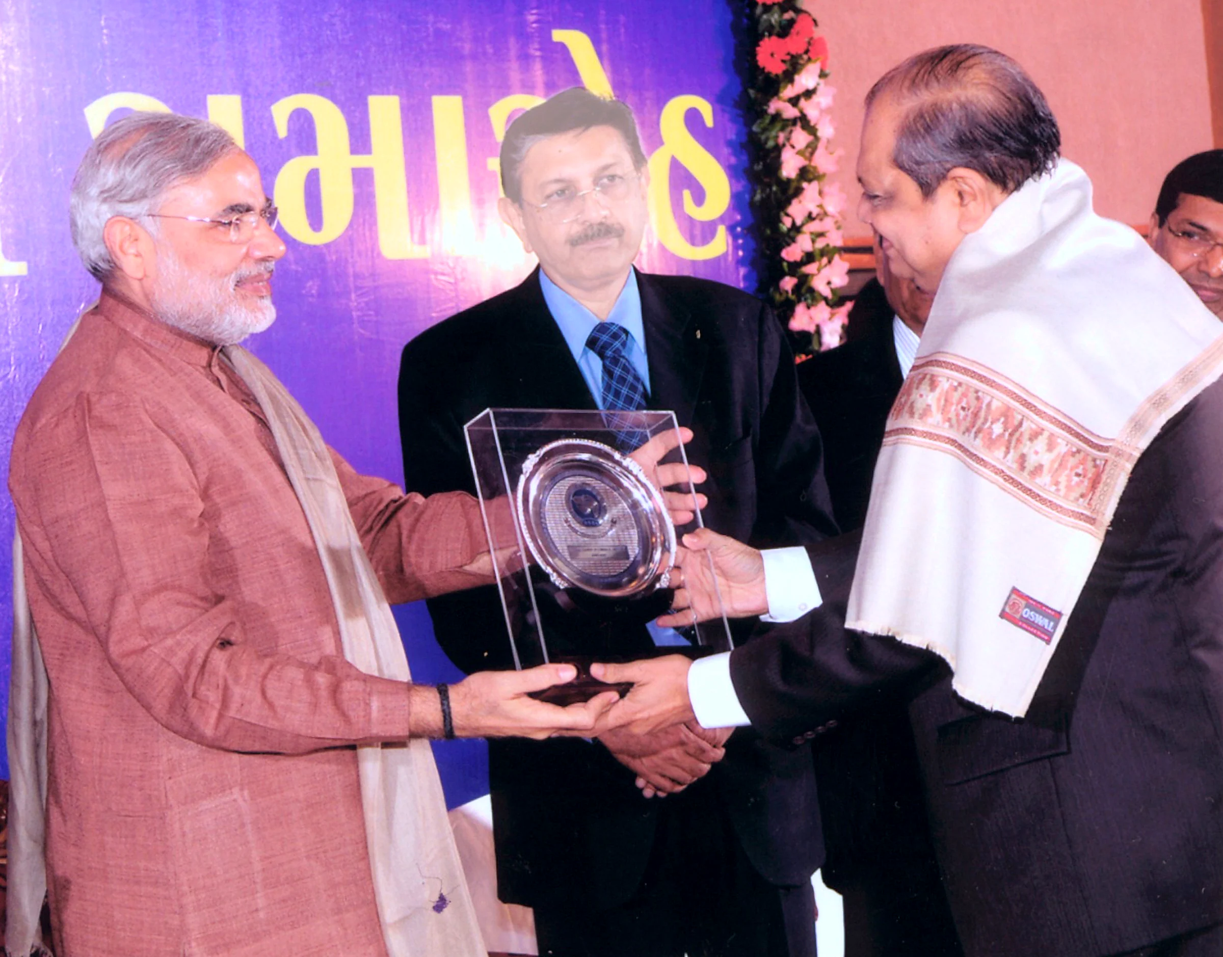 Narendra Modi presenting an award in a glass case to a man in a ceremonial shawl, under a Gujarati banner.