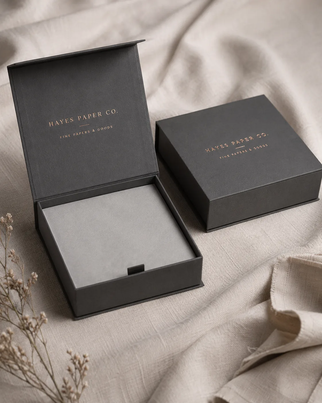 Two charcoal hinge lid cartons on a linen ground, one standing open on its hinge and one closed beside it.