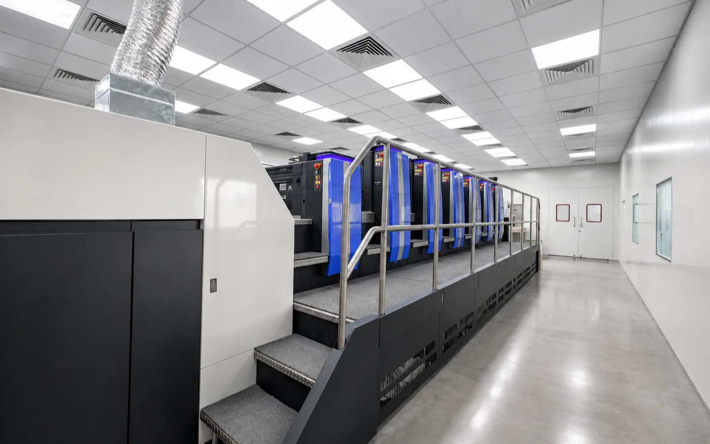The Komori Lithrone GL-637 six color offset press standing in a white walled press hall, its blue printing units in a row behind the access stair.