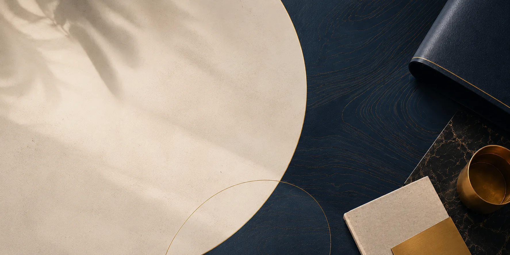 A round cream table mat with a fine gold rim on a dark navy wood table, seen from above, with a rolled navy mat beside it.