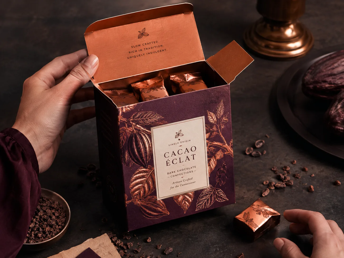 A deep aubergine chocolate carton in copper foil held open, the printed inside of the lid and the foil wrapped confections standing in it both in frame.