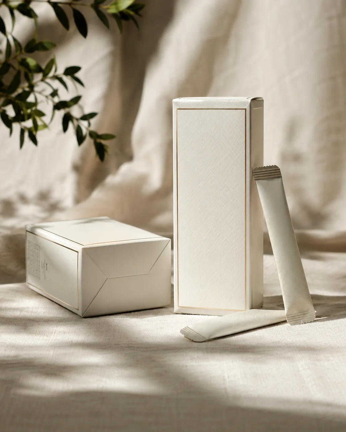 A cream seal end carton standing on linen beside a second laid flat to show its glued end, two stick pack sachets leaning against them.