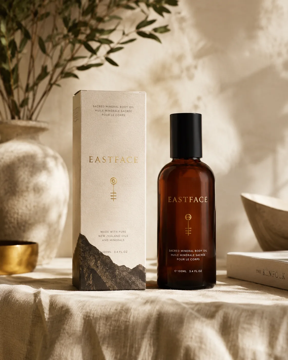 A cream straight tuck end carton standing on linen beside the amber body oil bottle it was drawn around, in directional daylight.