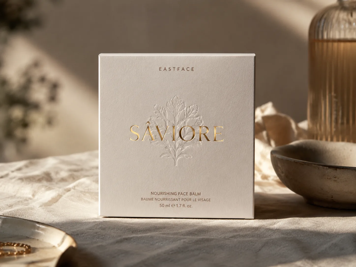 A square cream carton for a face balm on linen, a gold foil wordmark and a blind embossed botanical catching the light across the front panel.