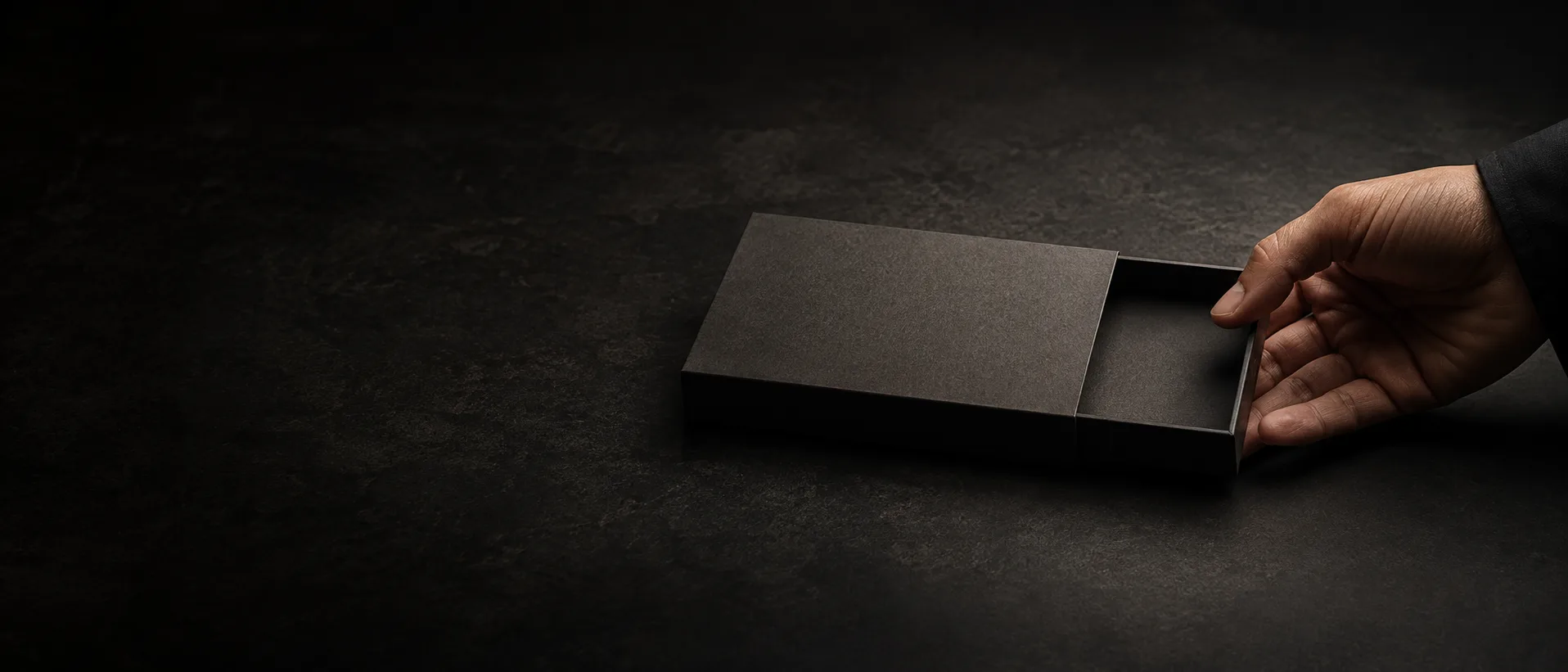 A hand drawing a tray out of its sleeve against a dark stone ground, the pack lit low from the right with the left of the frame in shadow.