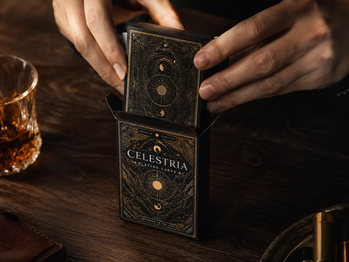 Two hands lowering a printed card deck into a black and gold tuck box on a dark table