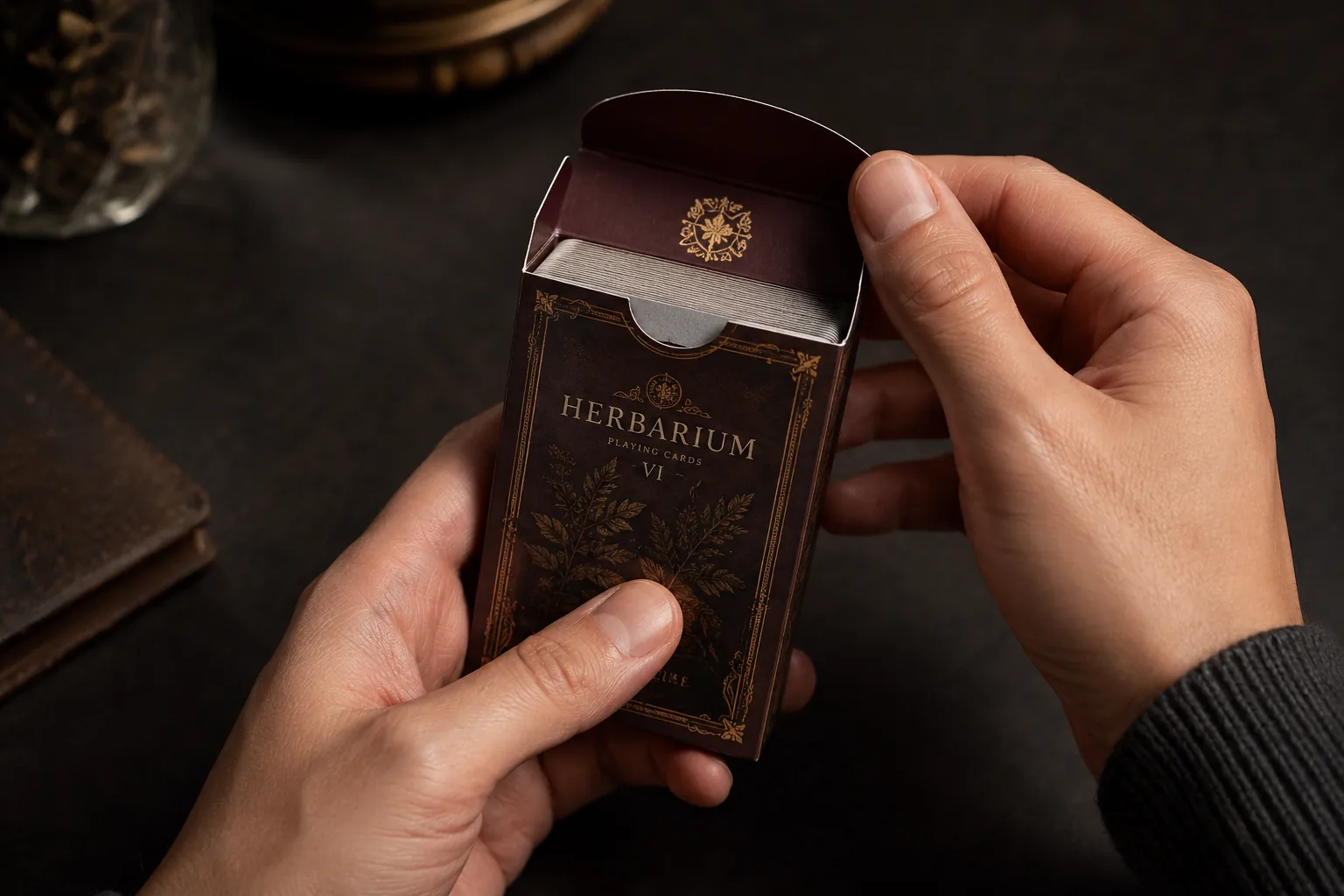 Two hands lifting the tuck flap of a printed deck box, the cards standing inside, on a dark table